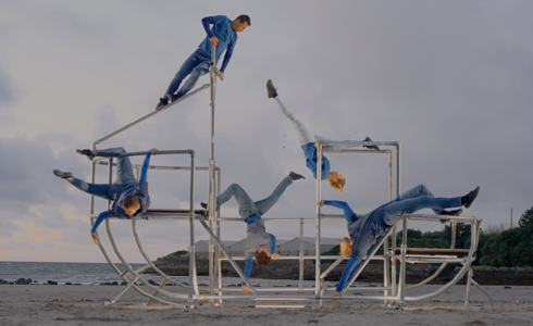 five dancers moving a metal shaped boat scaffolding structure on a beach. All wearing blue and grey outfits