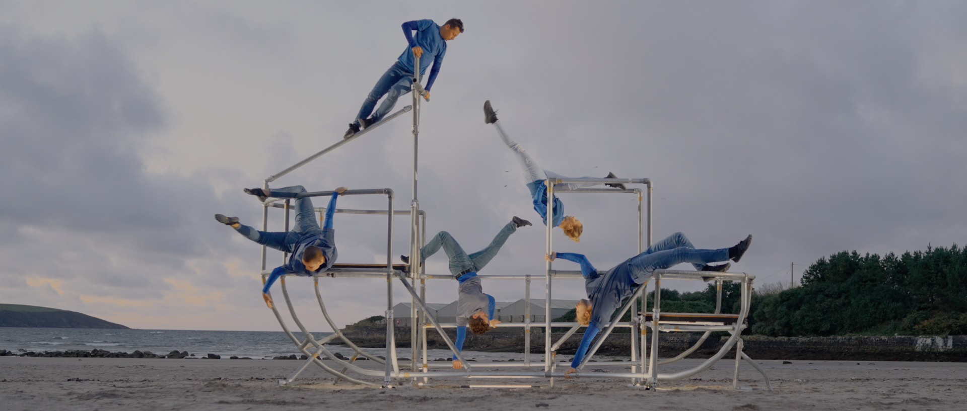 five dancers moving a metal shaped boat scaffolding structure on a beach. All wearing blue and grey outfits
