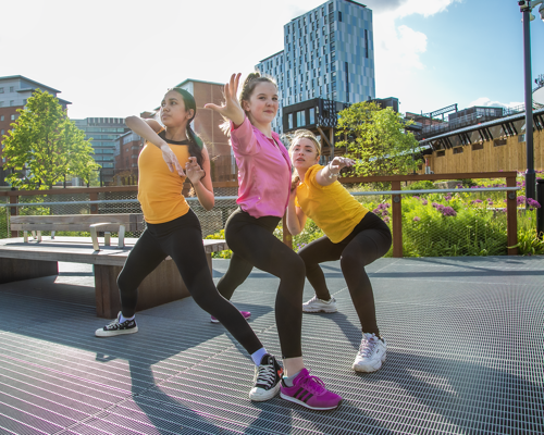 3 young female dancers infront of Machester skyline reaching out towards the camera. two girls wearing yellow tops black leggings and one girl wearing pink top black leggings 