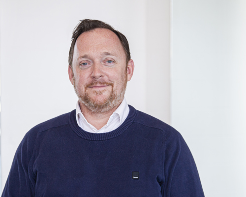 Headshot of Andrew Hurst. White male with light hair and beard smiling at camera, wearing blue jumper 