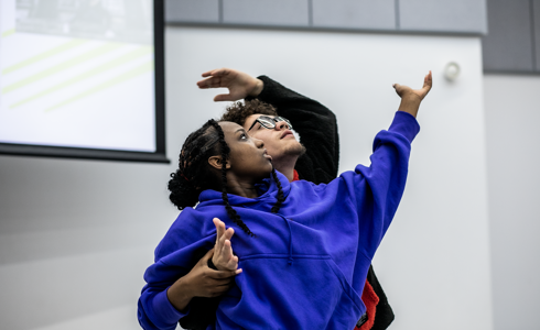 Two dancers, one global majority female with one arm high wearing purple hoodie and a male global majority dancer hugged around female with arm reaching to the top right wearing wholly black jumper. 