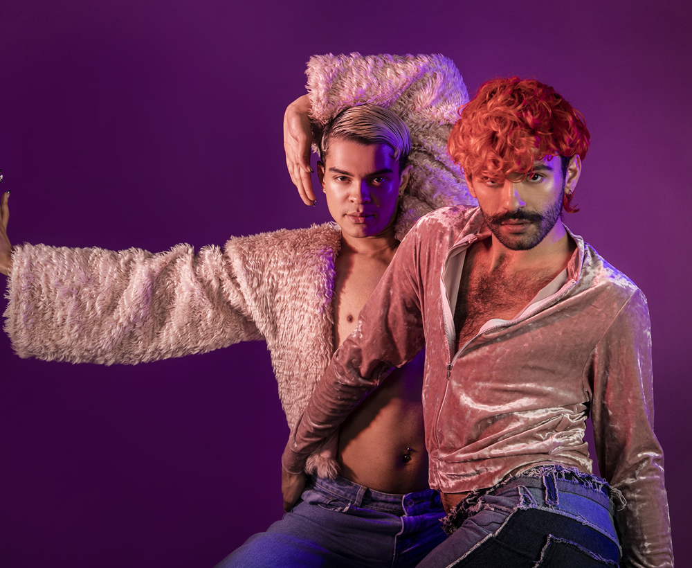 Two global majority LGBTQ+ male voguing dancers. Wearing pink tops and blue jeans on purple studio background. 