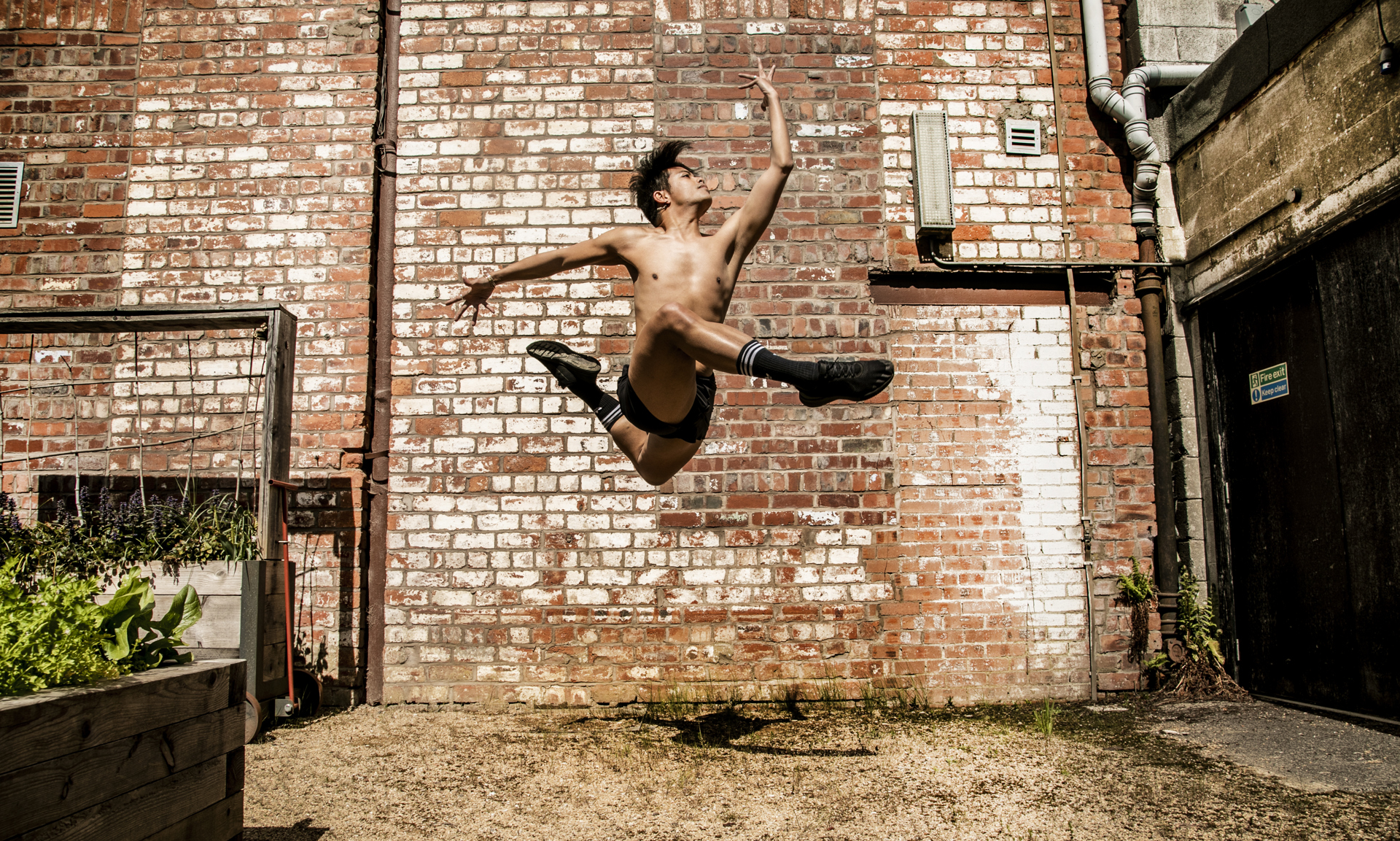 Global majority male dancer jumping high with legs tucked in a running shape, with one arm bent infront of him hand facing the sky and the other arm bent behind him. Wearing no t-shirt and black shorts with black trainers in front of red brick wall Global majority male dancer jumping high with legs tucked in a running shape, with one arm bent infront of him hand facing the sky and the other arm bent behind him. Wearing no t-shirt and black shorts with black trainers in front of red brick wall
