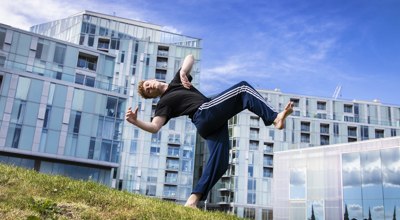 yong white male red haired dancer wearing blue tracksuit bottoms and black t-shirt. leaning back in a running man position on grassey hill in front of Trinity Laban University building 