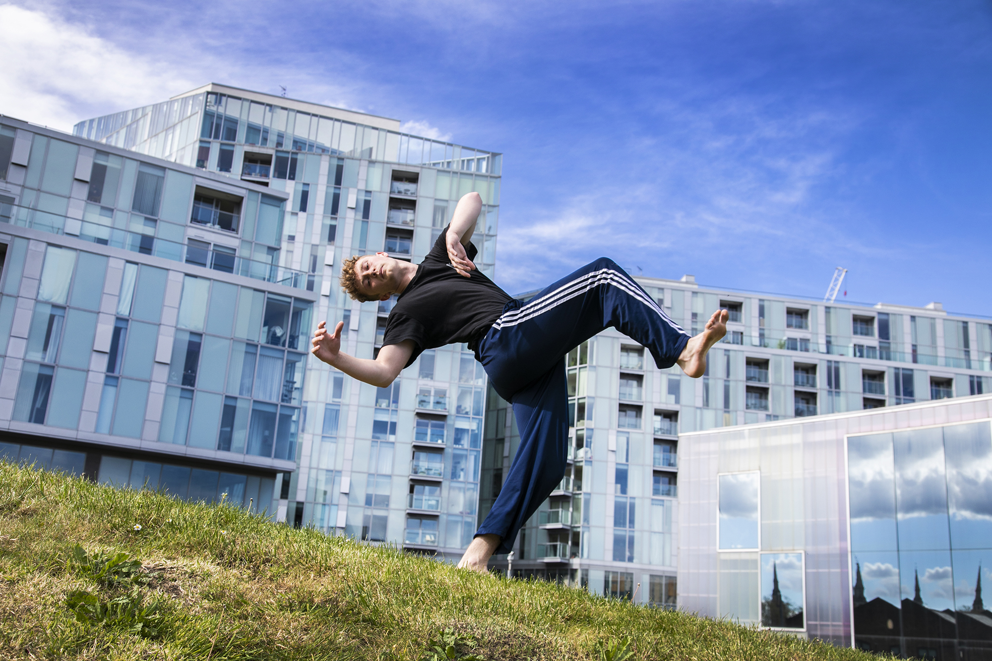 yong white male red haired dancer wearing blue tracksuit bottoms and black t-shirt. leaning back in a running man position on grassey hill in front of Trinity Laban University building  yong white male red haired dancer wearing blue tracksuit bottoms and black t-shirt. leaning back in a running man position on grassey hill in front of Trinity Laban University building