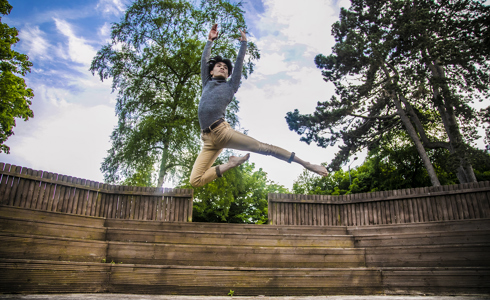 global majority male dancer jumping high with arms above head. Wearing grey long sleve tshirt and beige trousers.  In front of soft focus trees and blue sky.