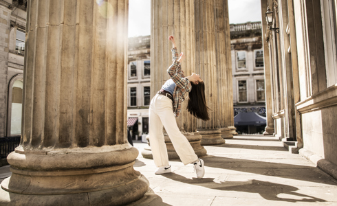 Young female dancer infront of Glasgow coucil building with large pillars. Legs lunched arms above the head. White female dance with long brown hair wearing checked shirt blue top and white trousers 