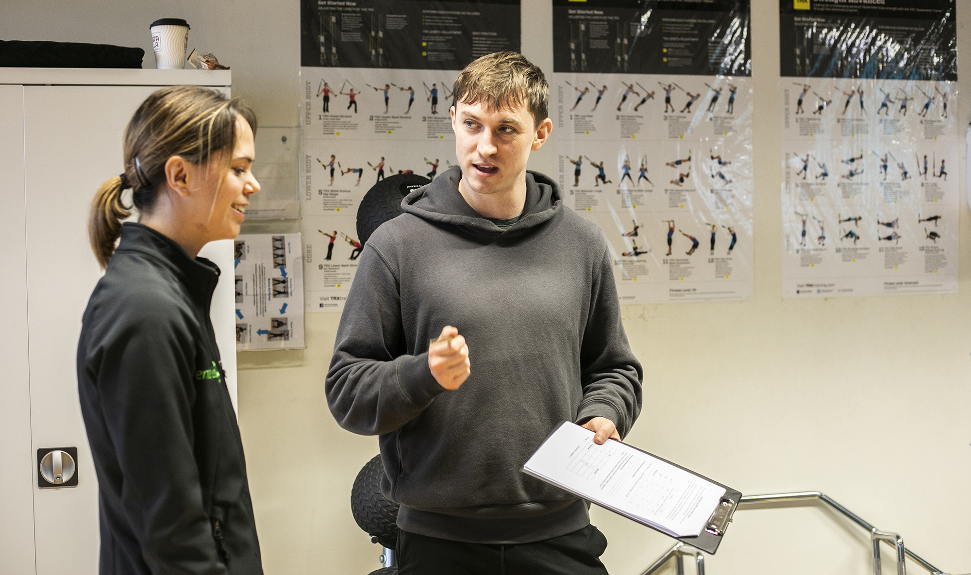 White male healthcare practitioner holding clipboard explainig something to white female in a healthcare environment with excersise posters in the background