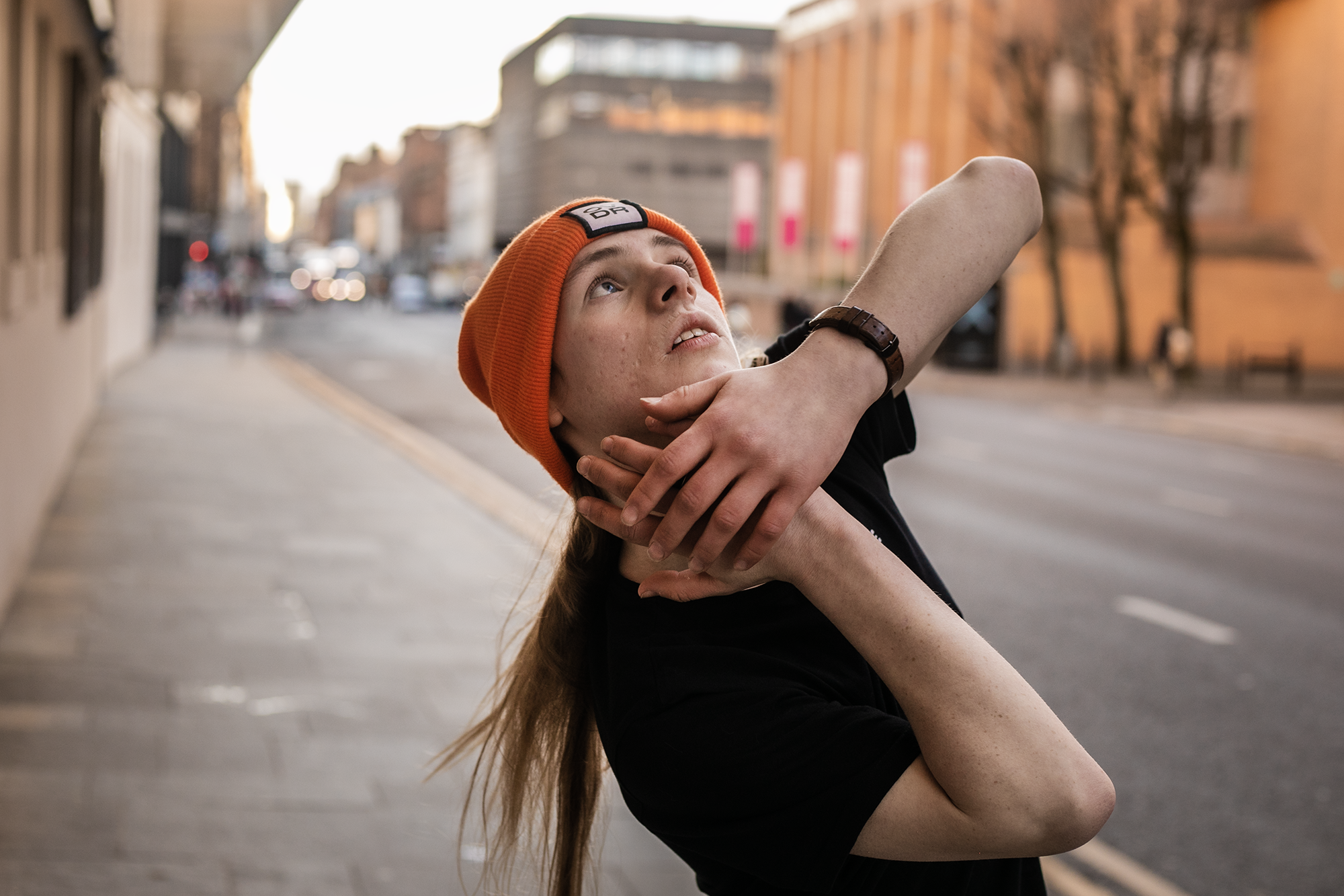 white male dancer with long blonde hair and orage beanie hat and black t shirt. On the street with hand around the face  white male dancer with long blonde hair and orage beanie hat and black t shirt. On the street with hand around the face