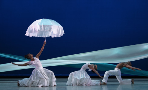 three global majority dancers on a blue stage with a blue ribbon graphic behind them. Female to the left lunging and leaning back holding up a white umbrella. Two male and female dancers on the right hand side lunging and arching back. All wearing white.