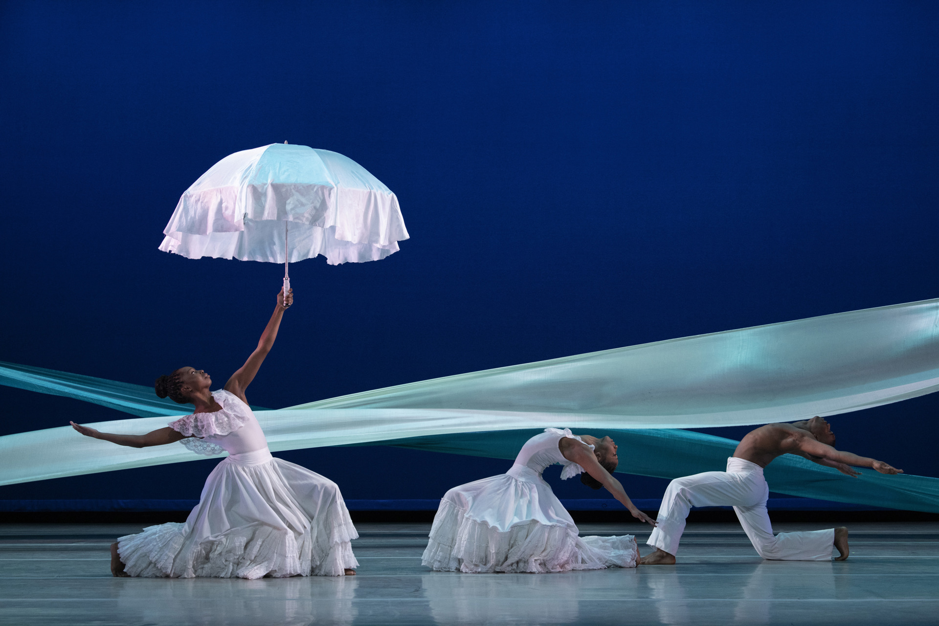 three global majority dancers on a blue stage with a blue ribbon graphic behind them. Female to the left lunging and leaning back holding up a white umbrella. Two male and female dancers on the right hand side lunging and arching back. All wearing white. three global majority dancers on a blue stage with a blue ribbon graphic behind them. Female to the left lunging and leaning back holding up a white umbrella. Two male and female dancers on the right hand side lunging and arching back. All wearing white.