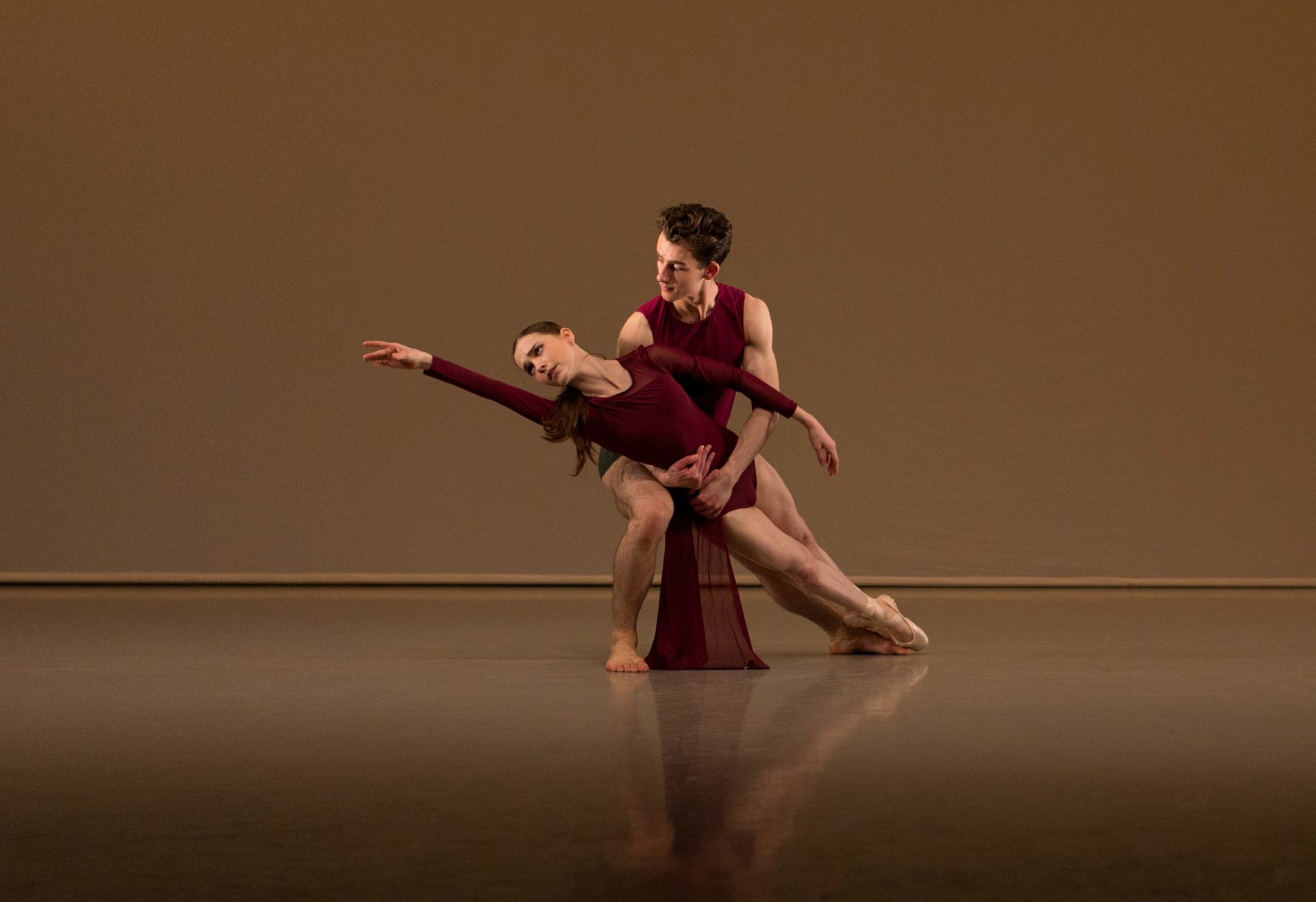 White male ballet dancer holding white female ballet dancer who is reaching to the upper left hand corner. Wearing red leotards. On a beige background White male ballet dancer holding white female ballet dancer who is reaching to the upper left hand corner. Wearing red leotards. On a beige background