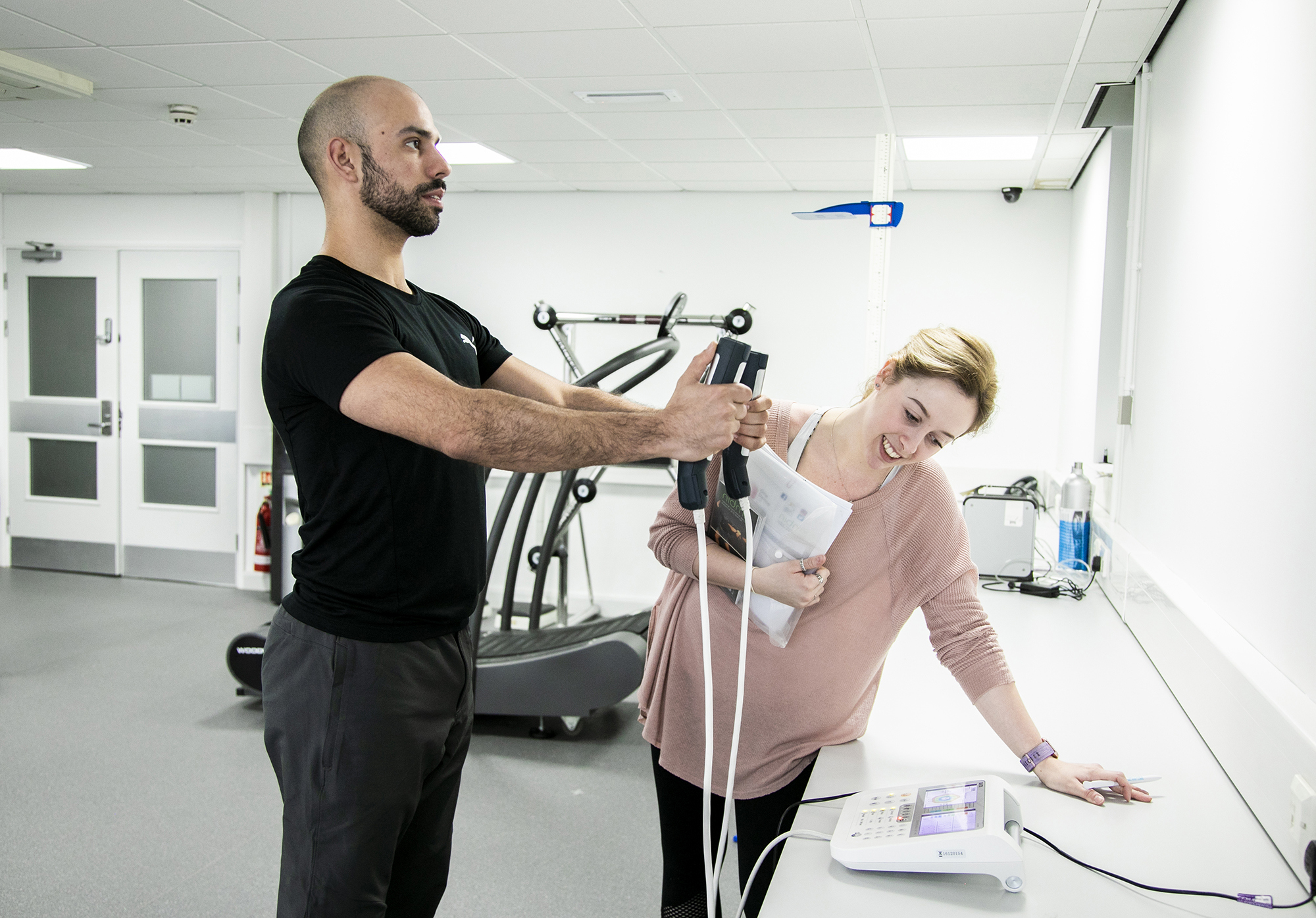 A male dancer is having his body composition measured by standing on a small scale with his arms outstretched in front of him. In either hand he is grasping a body-composition measurement device which are each connected to the scale with white cables. A white female screening assistant is leant over a white desk observing the monitor, and holding some papers. They are in a white and grey dance laboratory, with gym equipment in the background.  A male dancer is having his body composition measured by standing on a small scale with his arms outstretched in front of him. In either hand he is grasping a body-composition measurement device which are each connected to the scale with white cables. A white female screening assistant is leant over a white desk observing the monitor, and holding some papers. They are in a white and grey dance laboratory, with gym equipment in the background.