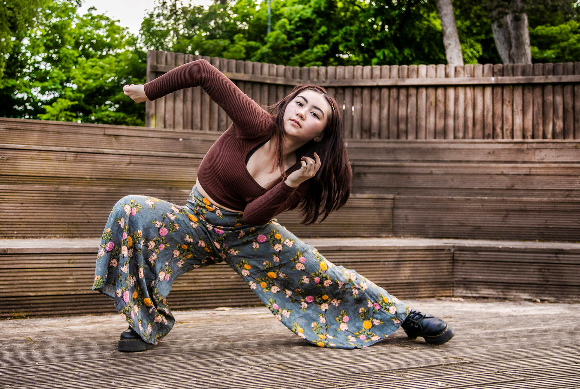 global majority female dancer lunging to the left with albow pointed in the air. In front of soft focus trees and wooden gate. Wearing brown long sleeved t-shirt and flowered flared trousers.  global majority female dancer lunging to the left with albow pointed in the air. In front of soft focus trees and wooden gate. Wearing brown long sleeved t-shirt and flowered flared trousers.