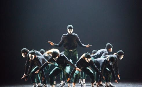 group of dancers on stage crouching down wearing white masks with one standing in the middle with arms stretched wide