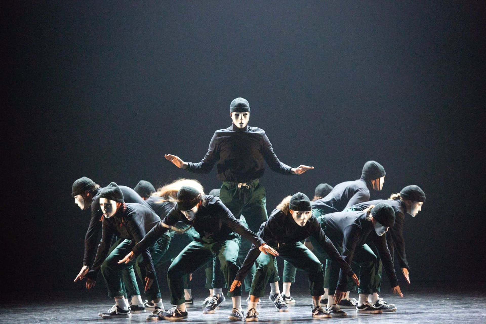 group of dancers on stage crouching down wearing white masks with one standing in the middle with arms stretched wide