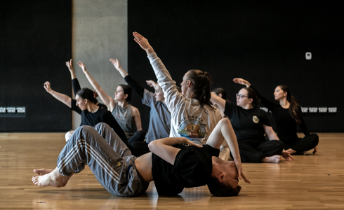 7 female dancers sitting crossed legged with one arm in the air. one male dancer infront laying on the floor with knees up arched back and hand infront of face. In a dance stucio with wooden floors and black walls 