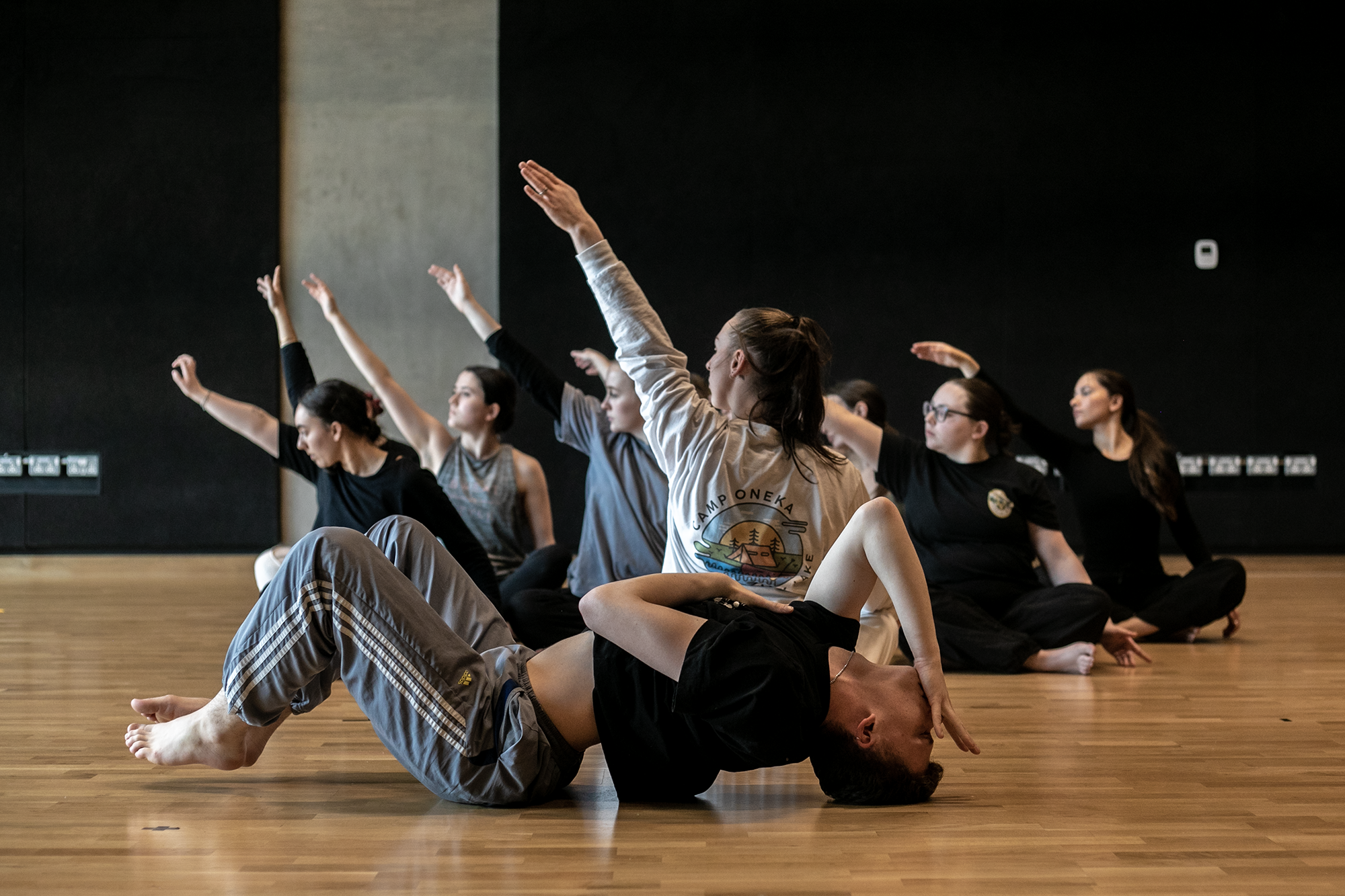 7 female dancers sitting crossed legged with one arm in the air. one male dancer infront laying on the floor with knees up arched back and hand infront of face. In a dance stucio with wooden floors and black walls  7 female dancers sitting crossed legged with one arm in the air. one male dancer infront laying on the floor with knees up arched back and hand infront of face. In a dance stucio with wooden floors and black walls