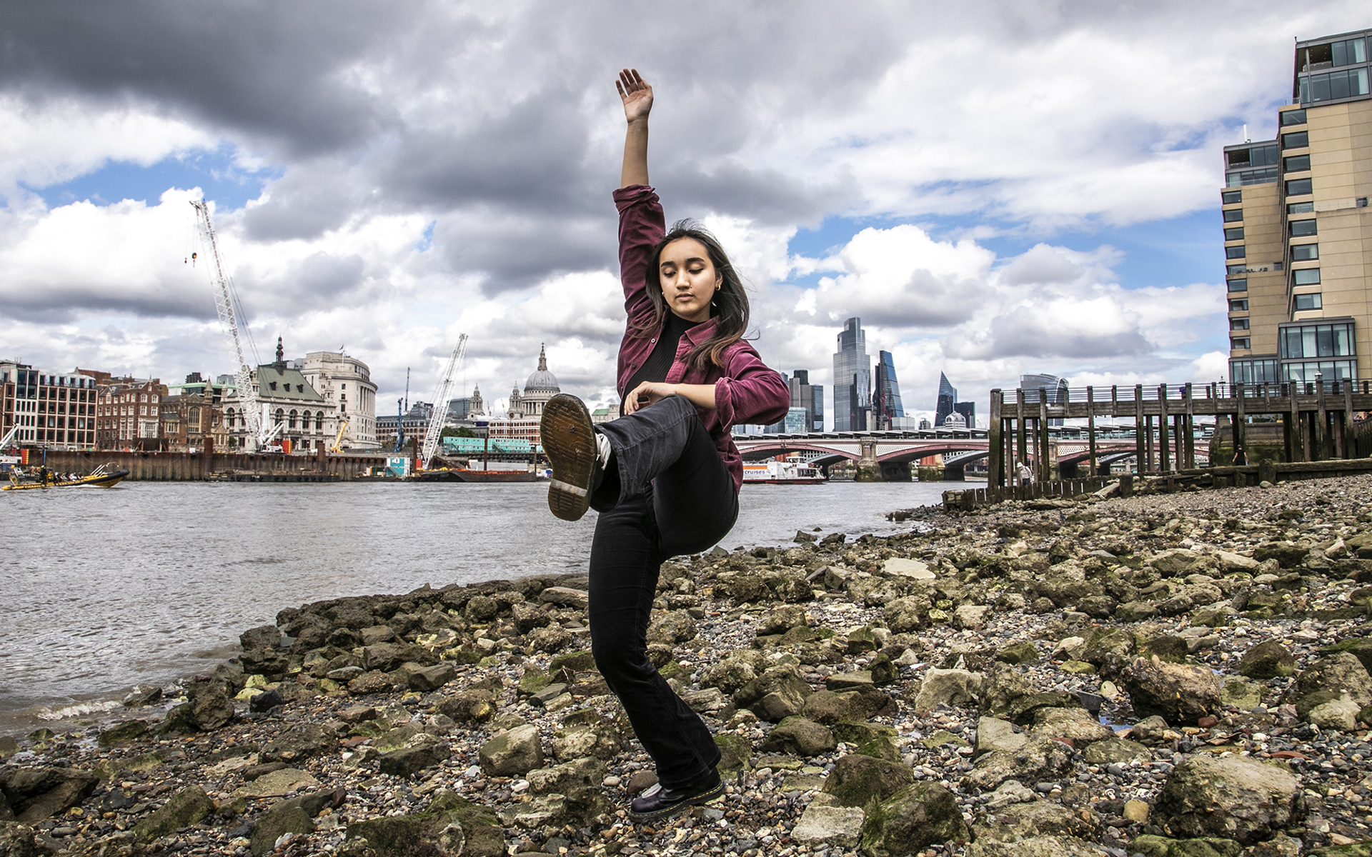 Female contemporary dancer with one leg risen and one arm above the head. Wearing black trousers and red open shirt standing with london beach skyline Female contemporary dancer with one leg risen and one arm above the head. Wearing black trousers and red open shirt standing with london beach skyline
