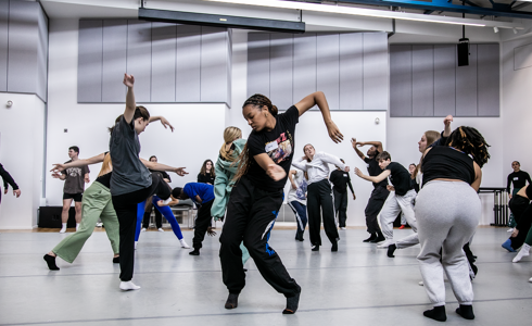 Young people dancing in a dance studio all interacting with each other. Main focus on global majority female dancer moving her arms to the right and swinging hair around.