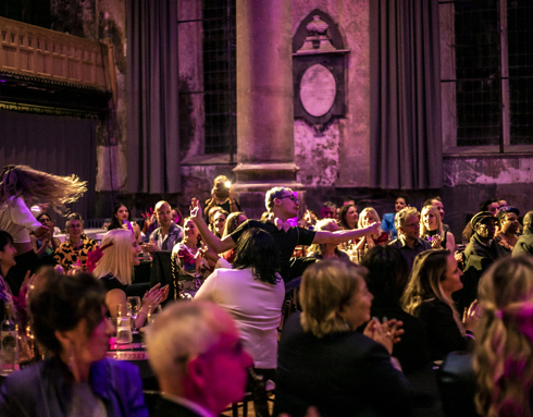 audience sitting down clapped as drag show dancers dance through the seats and tables. Large church venue is lit pink audience sitting down clapped as drag show dancers dance through the seats and tables. Large church venue is lit pink
