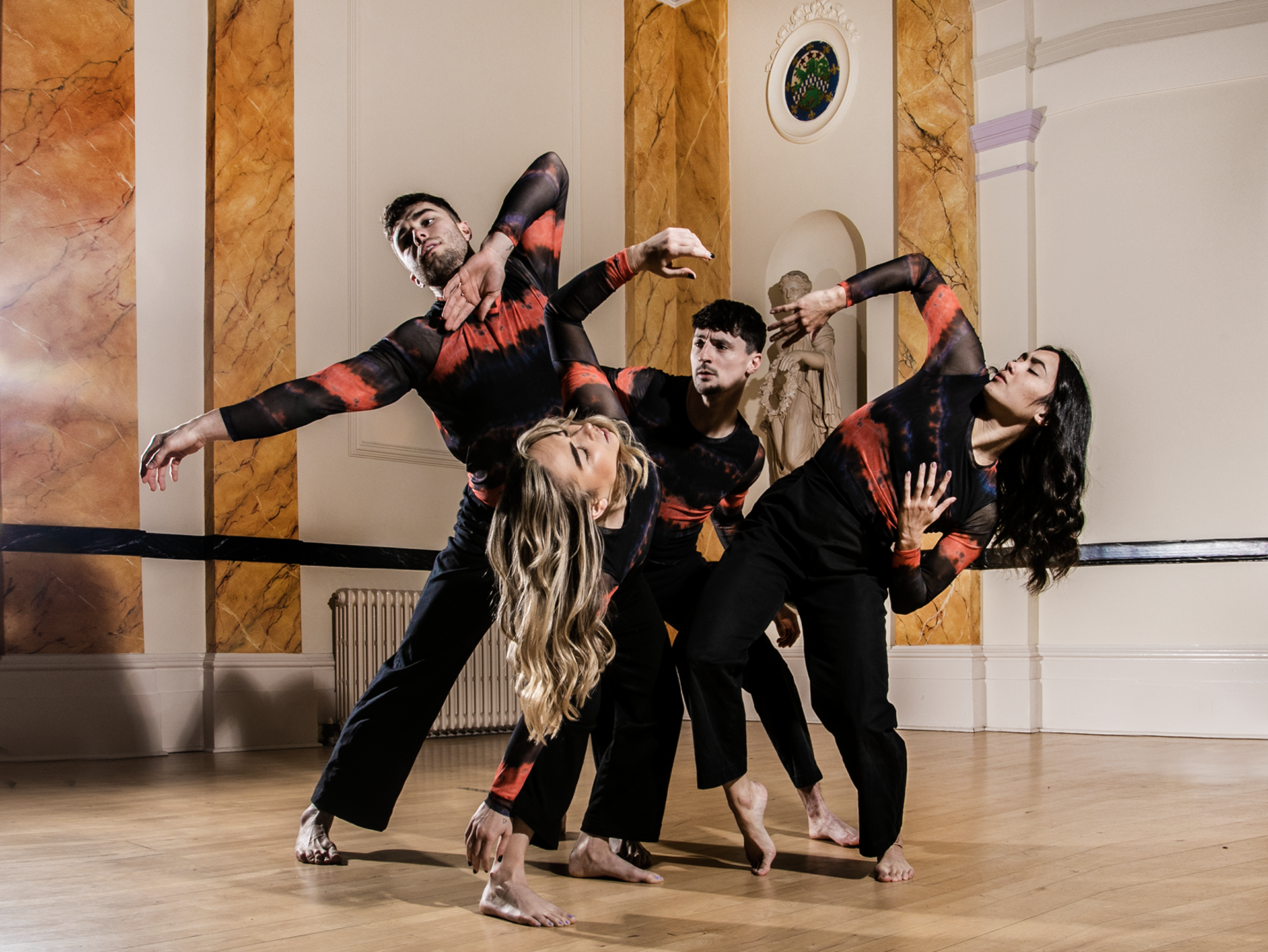 Four dancers intertwined with arms twisted around eachother in a museum building. two white male dancers 2 white female dancers all wearing black and red long sleve tops and trousers. Four dancers intertwined with arms twisted around eachother in a museum building. two white male dancers 2 white female dancers all wearing black and red long sleve tops and trousers.