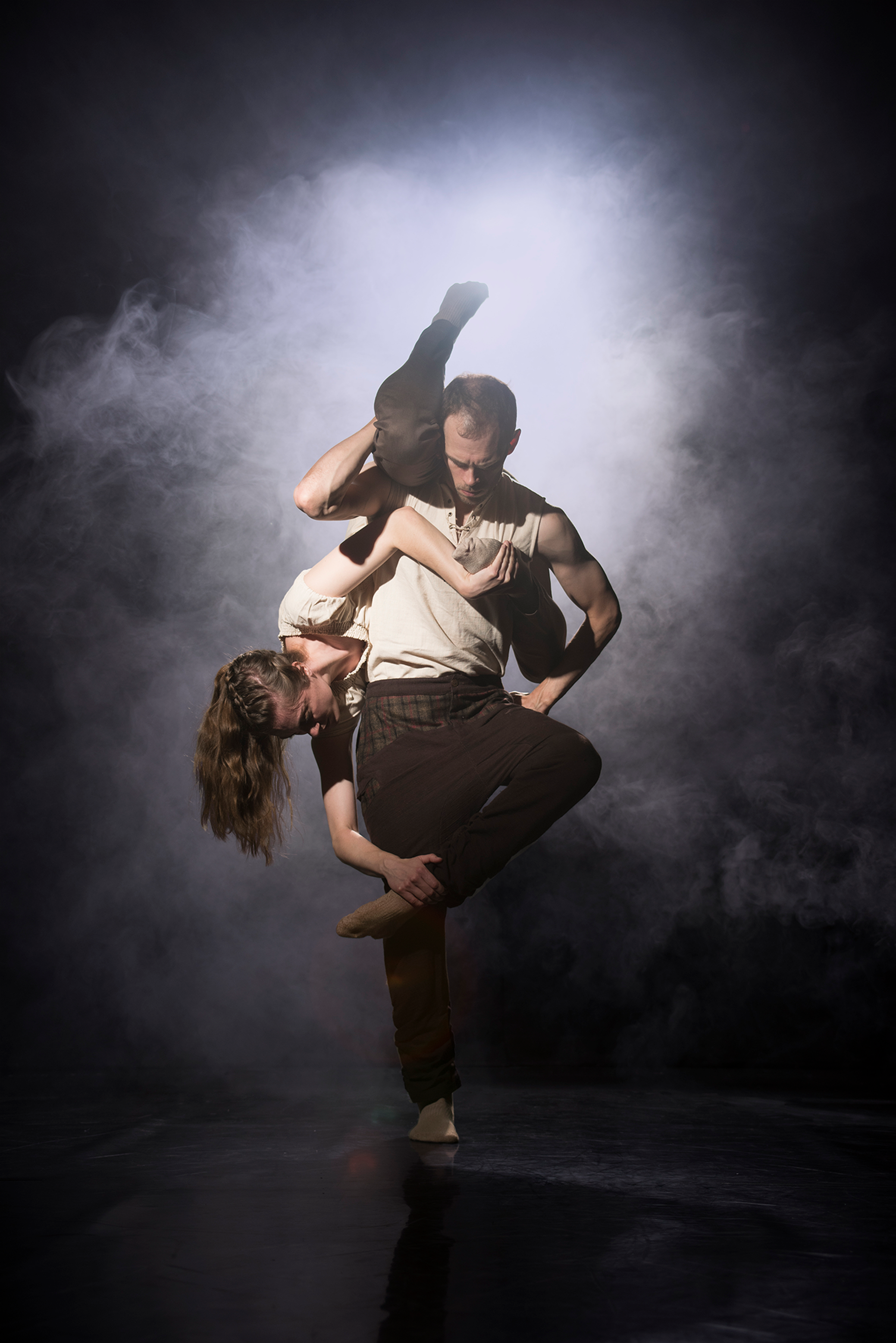 white male dancer standing straight with a white female dancer upside down wrapped around his body, both wearing white tops and black trousers on a smokey dark stage