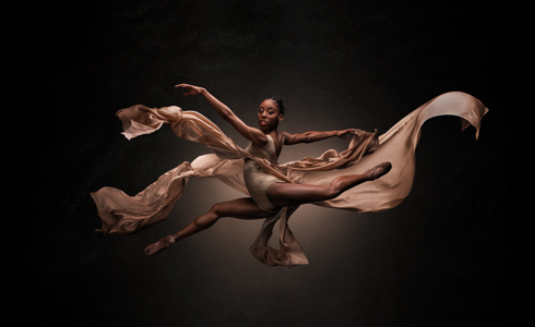 Global majority female dancer in a splits jump with arms out to the side and face towards the camera. Wearing nude coloured leotard with floating skirt