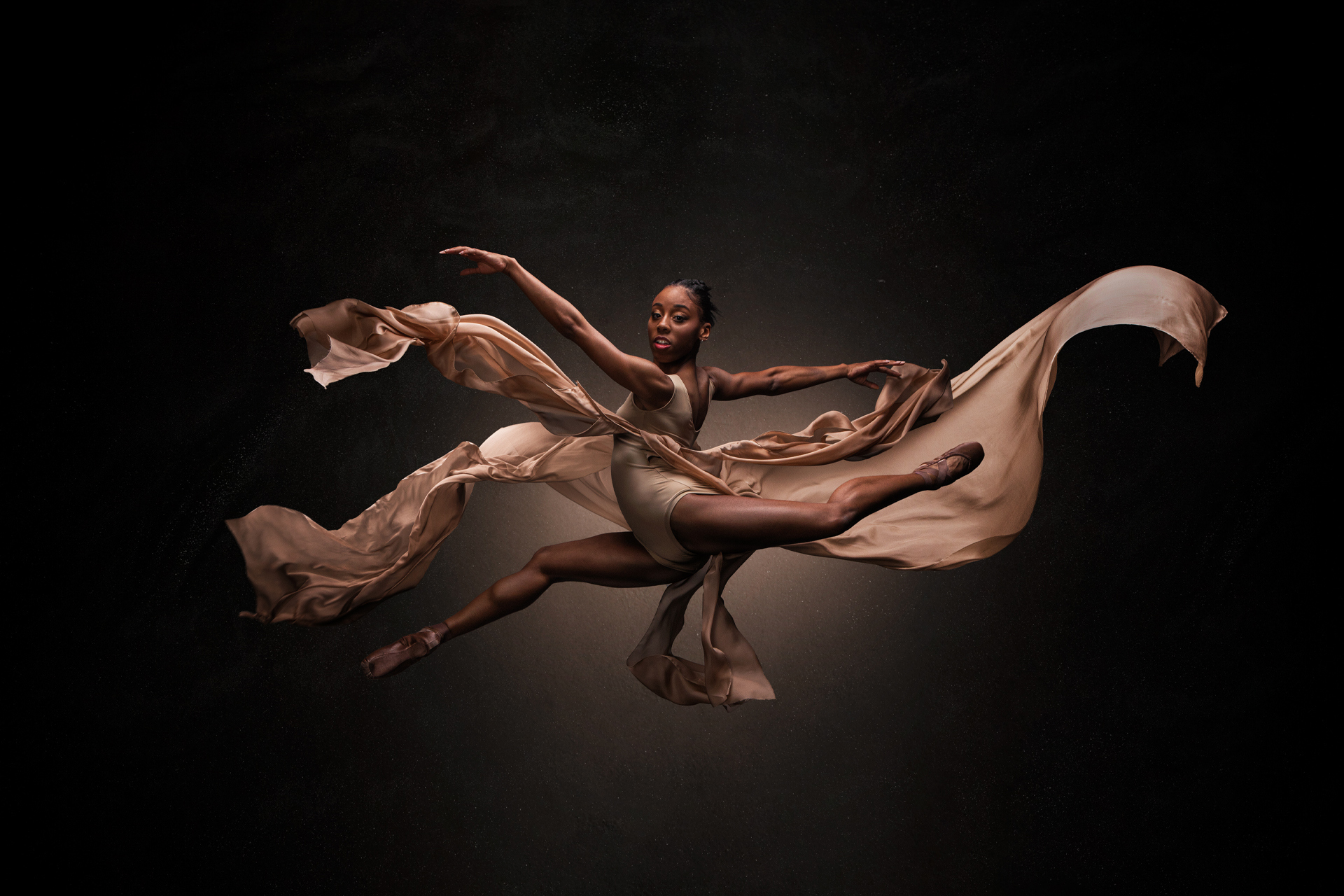 Global majority female dancer in a splits jump with arms out to the side and face towards the camera. Wearing nude coloured leotard with floating skirt