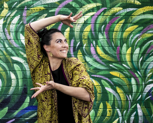 Global majority female dancer with long black hair smiling with arms framing the face. In front of green colourful graffiti wall. Wearing green and purple kaftan. 