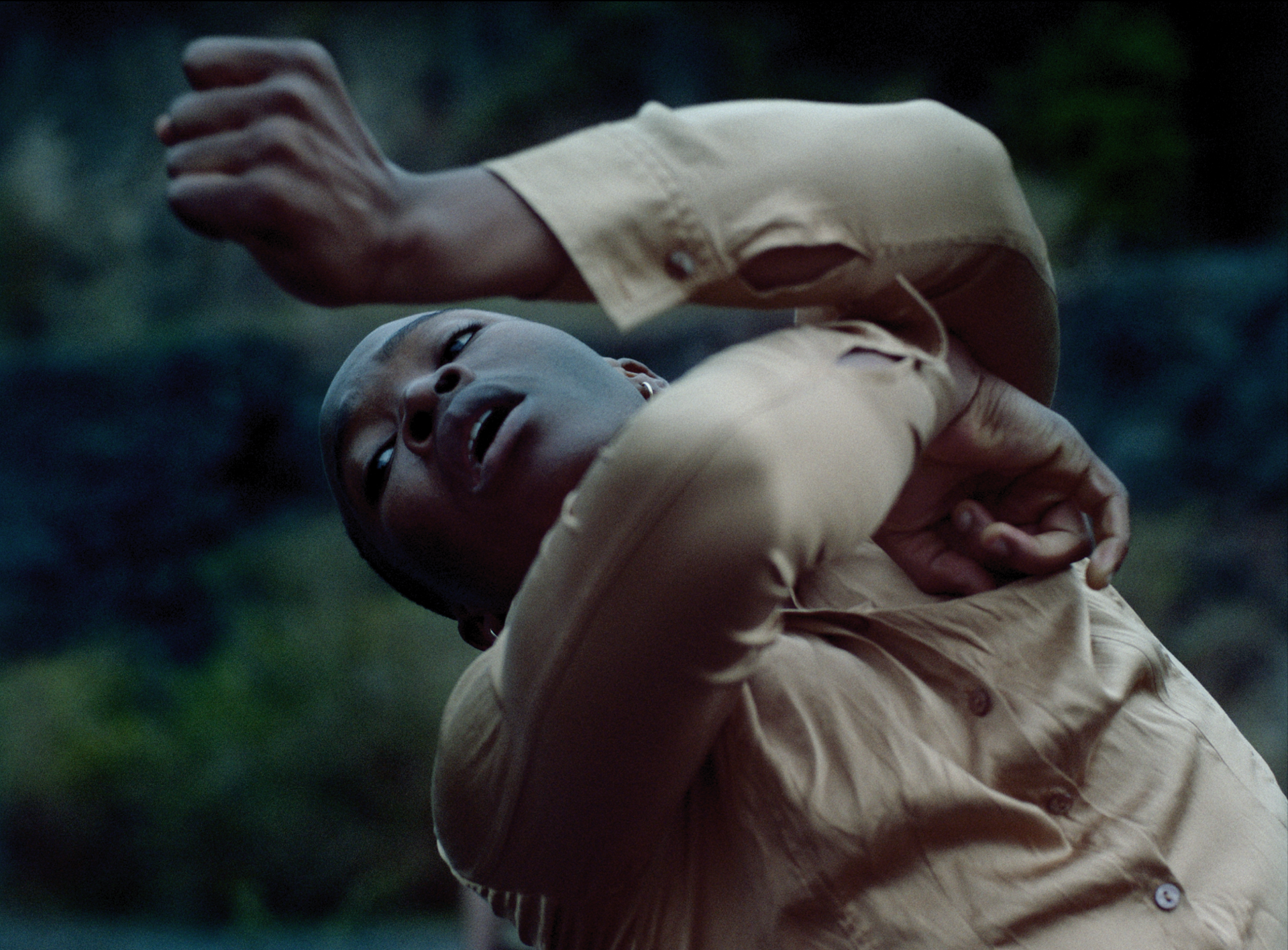 Close up of global majority dancer moving arms around the face with eyes up to the top left corner of the image, wearing muted clothes in front of dark wall Close up of global majority dancer moving arms around the face with eyes up to the top left corner of the image, wearing muted clothes in front of dark wall