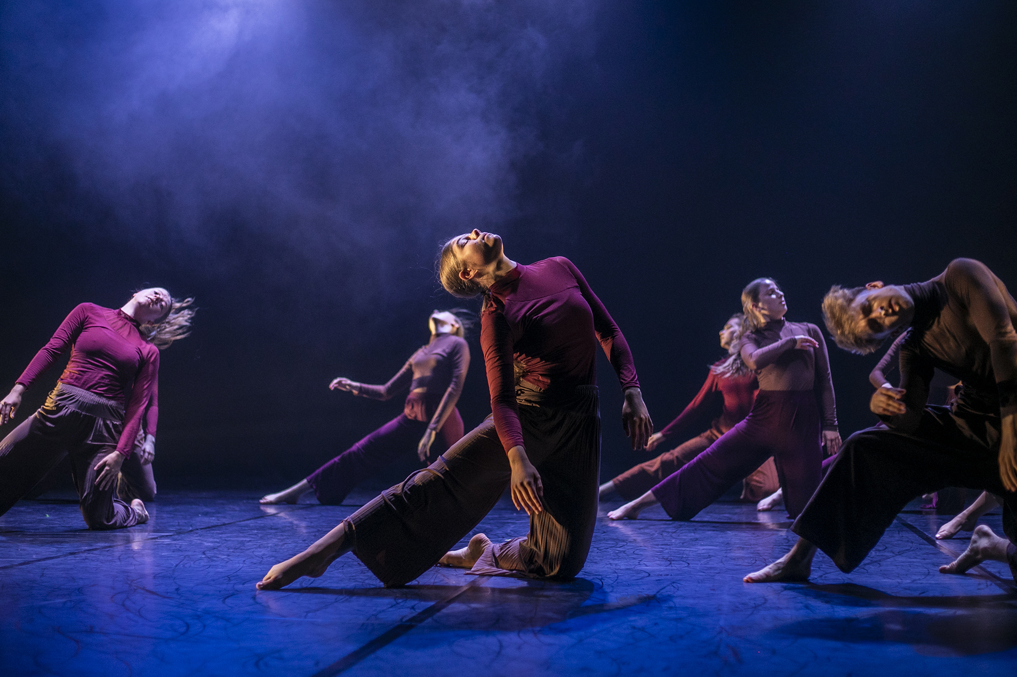 6 female dancers performing on the U.Dance stage wearing long sleve red tops and baggy brown trousers. All staners on one know with the other leg straight to the left with heads titled to the sky. Stage lighting is dark with blue fog and floor. 