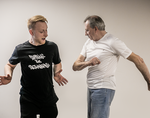 Two men body poppin dancing together. One young blonde white male dance teacher wearing all black, the other an older male grey haired dance student wearing blue jeans and white top. 