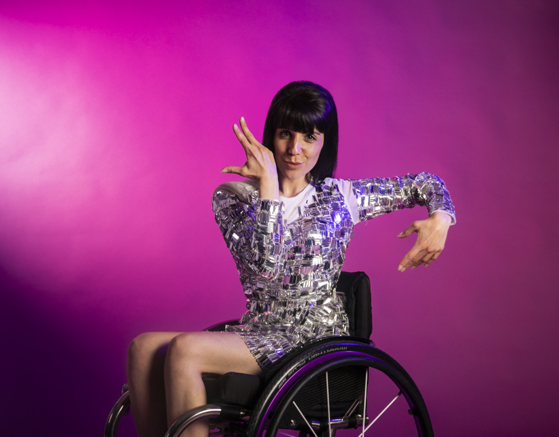 white brunette female dancer in a wheelchair wearing silver sparkly dress. Both hands in salsa movement around her face. On a pink photo backdrop white brunette female dancer in a wheelchair wearing silver sparkly dress. Both hands in salsa movement around her face. On a pink photo backdrop
