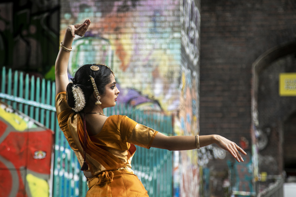 Back of a South Asian dancer dressed in traditional orange dress with hair jewellery. In front of colourful graffiti walls. One arm above her head and one arm stretched out. 