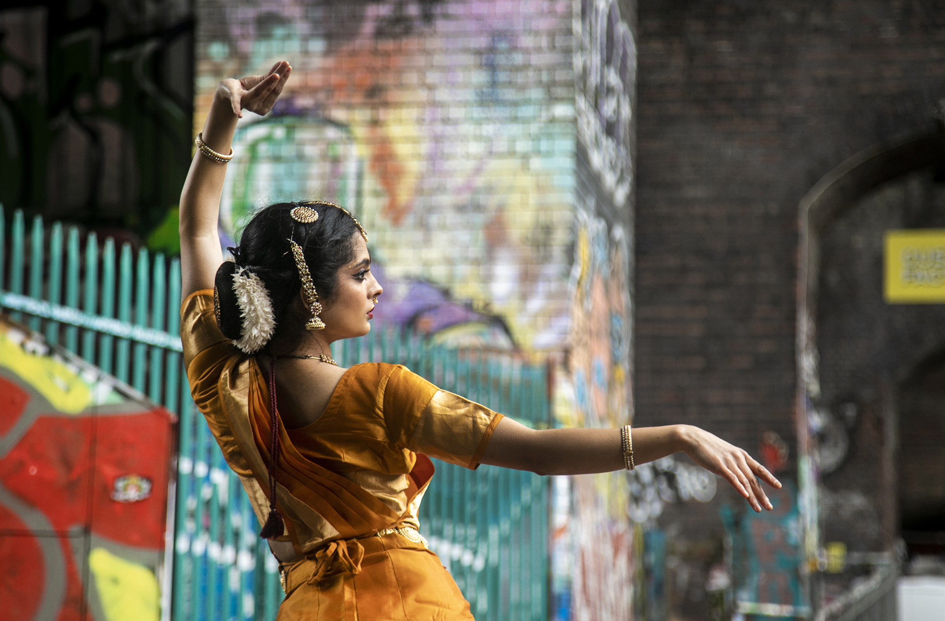 Back of a South Asian dancer dressed in traditional orange dress with hair jewellery. In front of colourful graffiti walls. One arm above her head and one arm stretched out. 