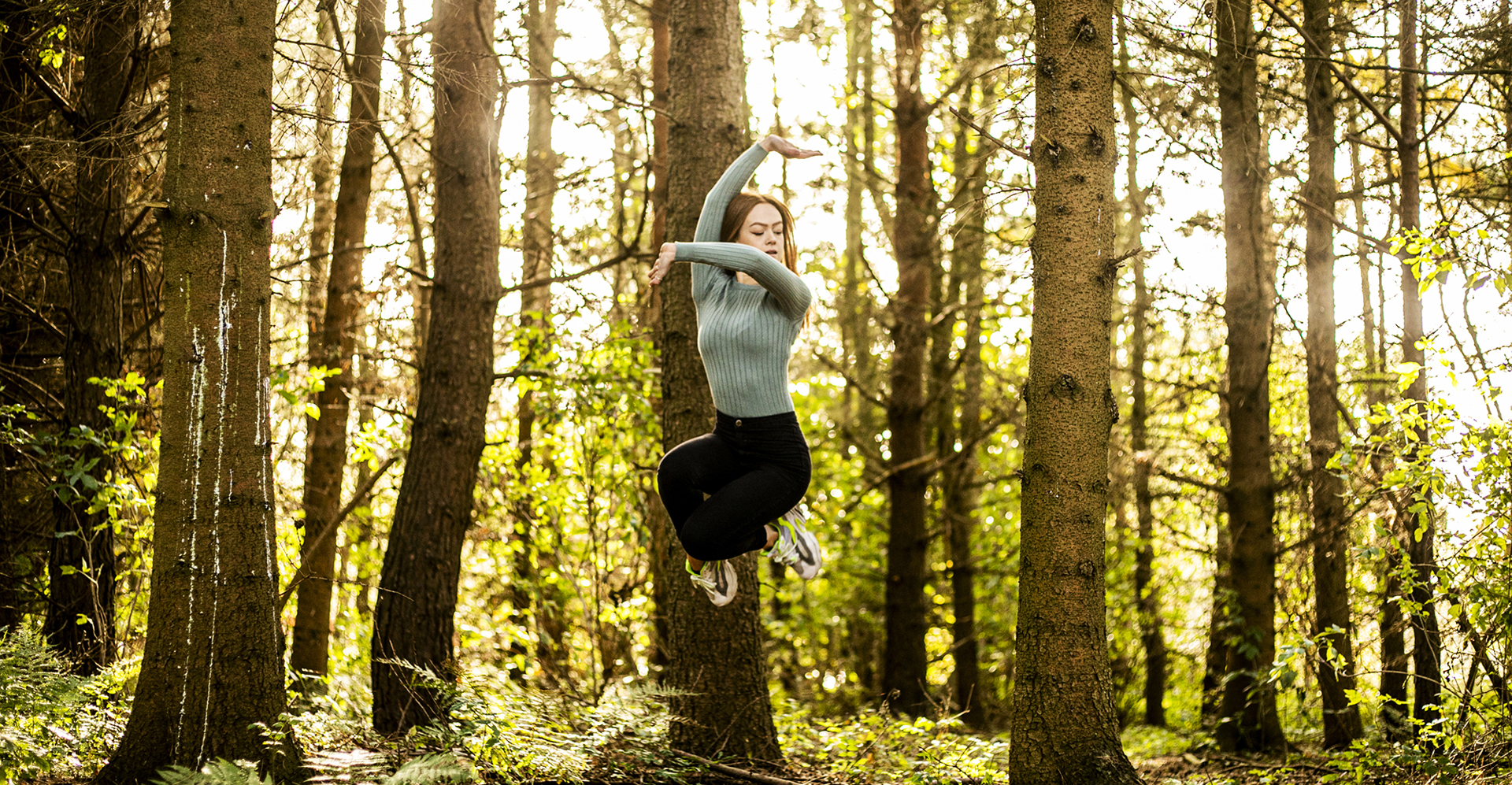 white female dancer jumping in the air with legs tucked and one arm up and the other across body. In front of woodland in golden light. white female dancer jumping in the air with legs tucked and one arm up and the other across body. In front of woodland in golden light.