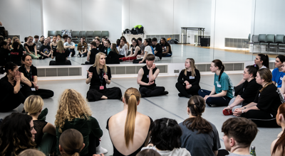 White female One Dance UK Staff member sitting on a dance studio floor talking to a large group sitting in a cirlce of young people. Mirrored walls behind them reflecting the group. 