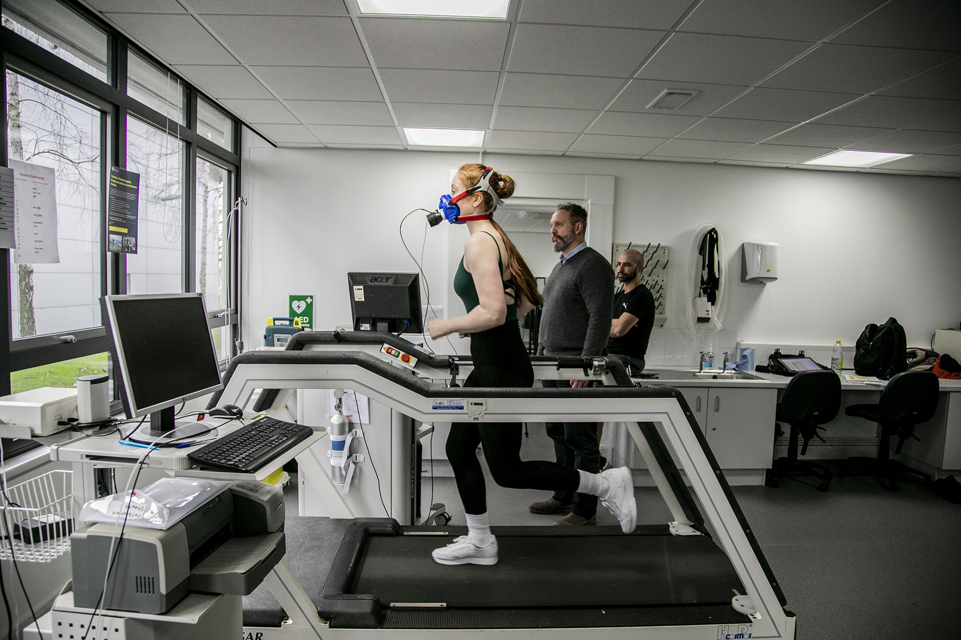 White female dancer with long red hair running on a treadmill wearing face-mask to measure cardiovascular fitness. White male physiologist looking over to observe stats on the machine. In a white laboratory setting with a window in the background White female dancer with long red hair running on a treadmill wearing face-mask to measure cardiovascular fitness. White male physiologist looking over to observe stats on the machine. In a white laboratory setting with a window in the background