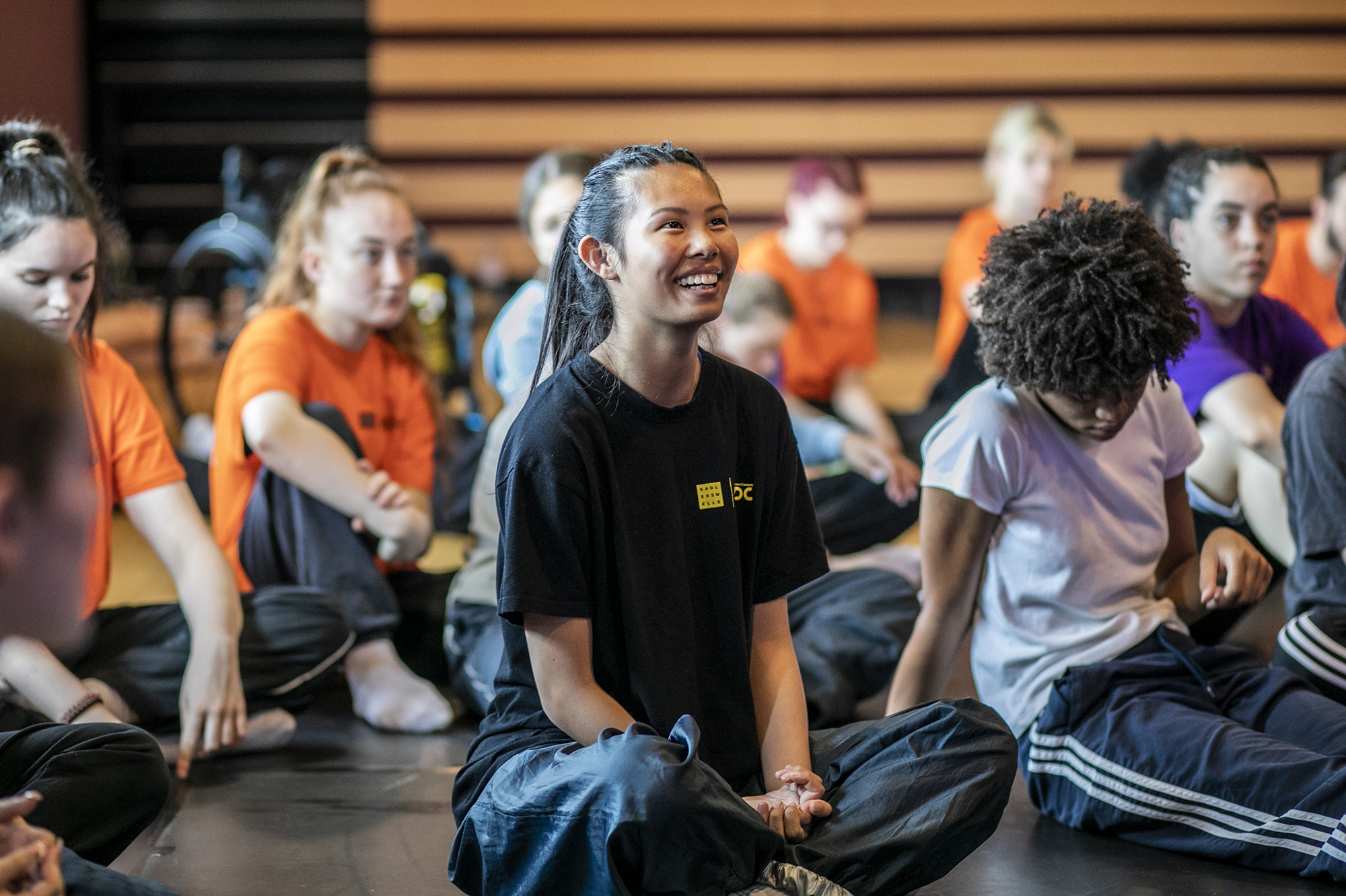 young dancers white and global majority, male and female, sitting crossed legged smiling and listening. 