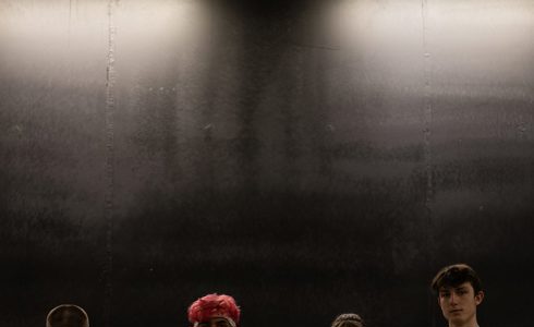 four young dancers wearing white tracksuits looking at the camera with dark walls behind them 