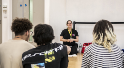 One Dance UK staff member sitting on knees talking to three students sitting in front of the camera in a dance studio.  One Dance UK staff member sitting on knees talking to three students sitting in front of the camera in a dance studio.