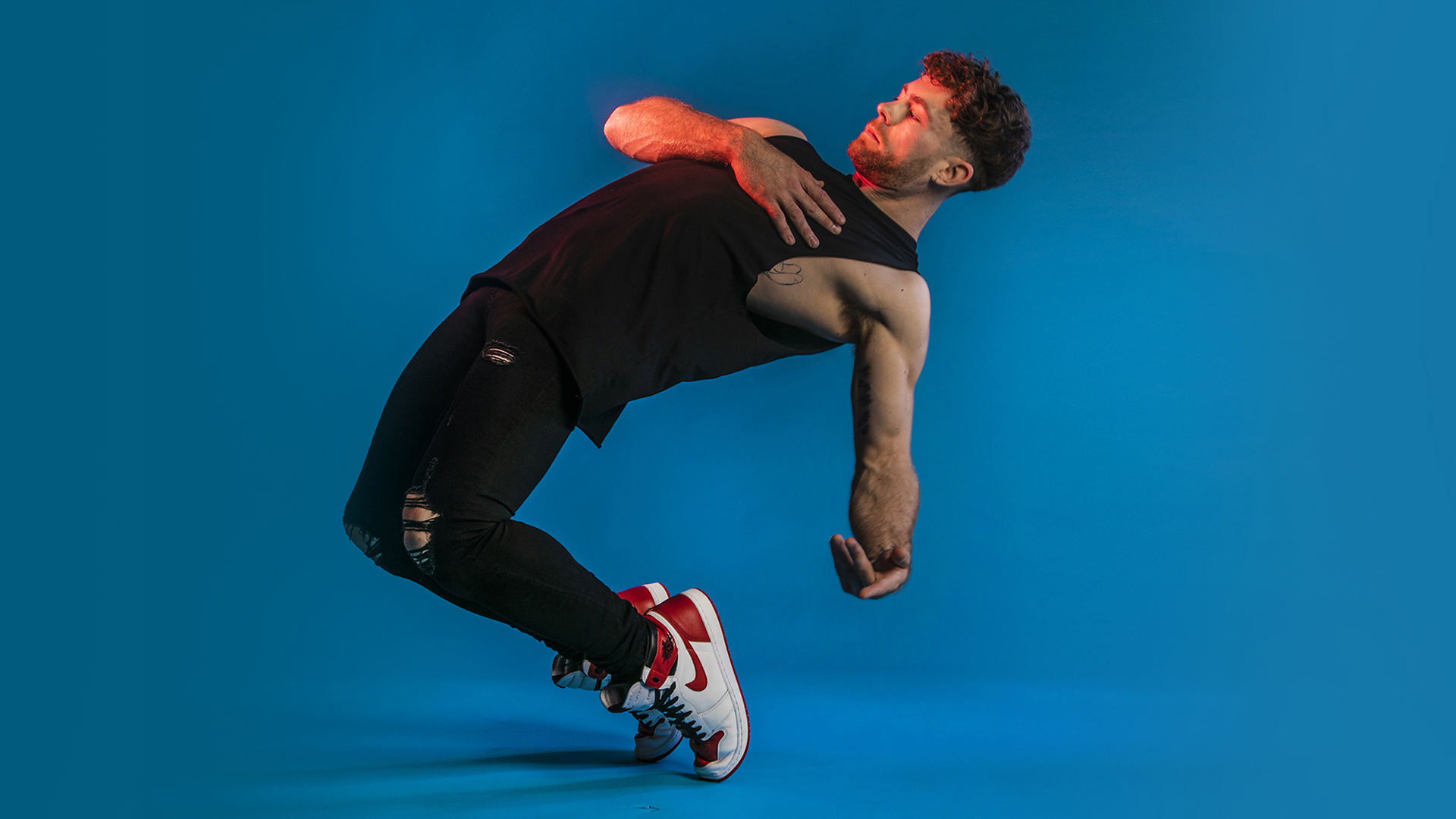 white male dancer leaning back on his tip toes. Black outfit and white trainers. On blue studio background. white male dancer leaning back on his tip toes. Black outfit and white trainers. On blue studio background.