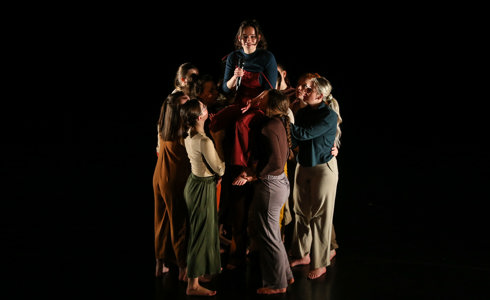 Group of female dancers huddled together lifting one person up in the middle on a dark stage