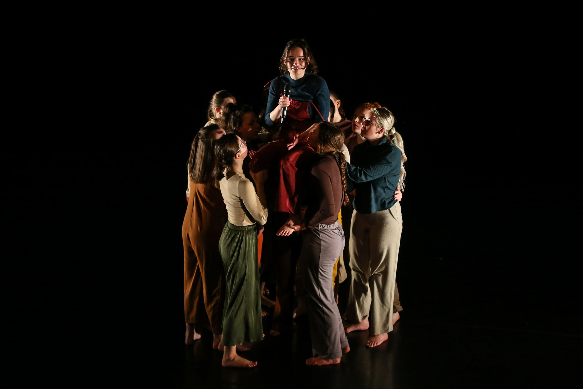 Group of female dancers huddled together lifting one person up in the middle on a dark stage