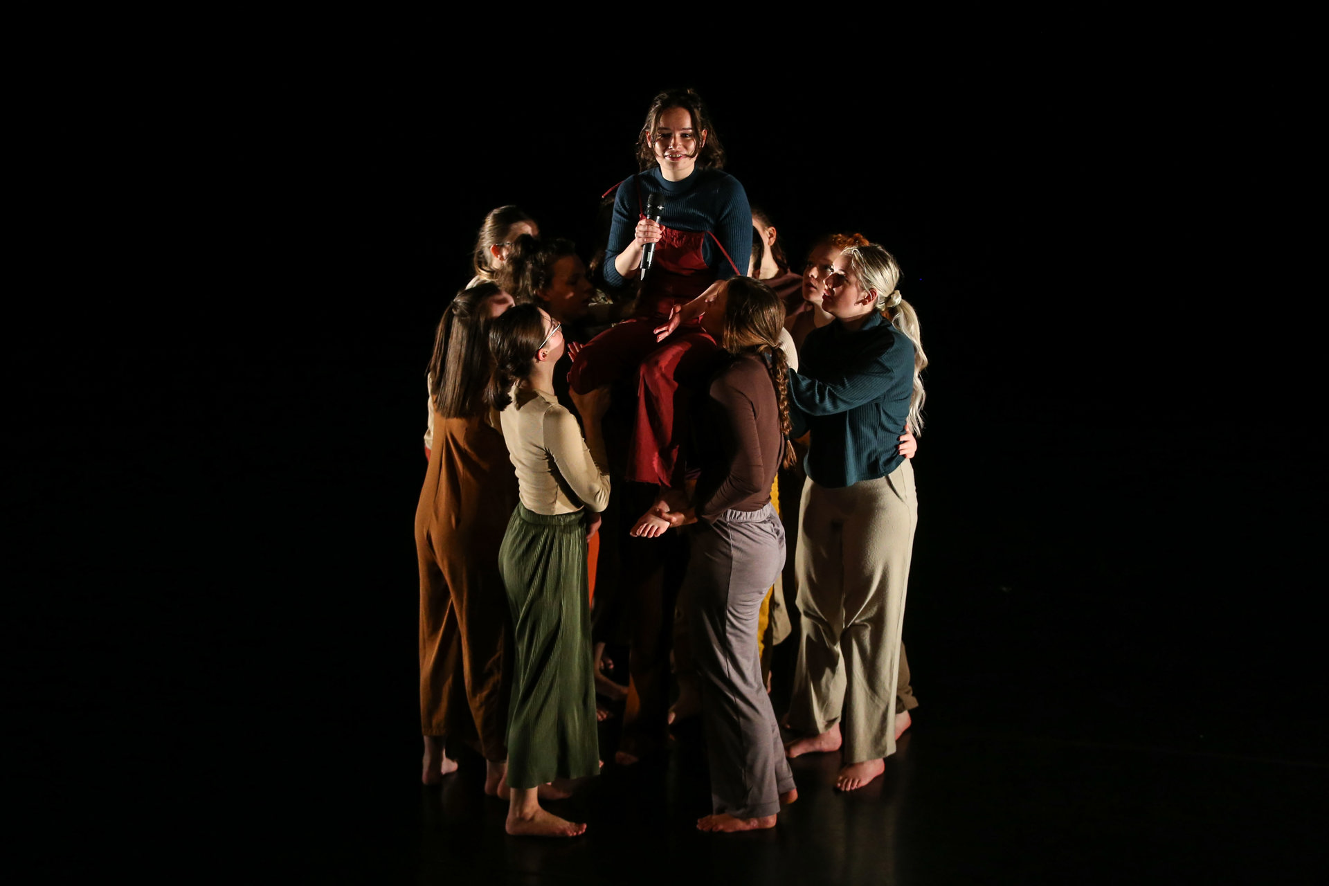 Group of female dancers huddled together lifting one person up in the middle on a dark stage Group of female dancers huddled together lifting one person up in the middle on a dark stage