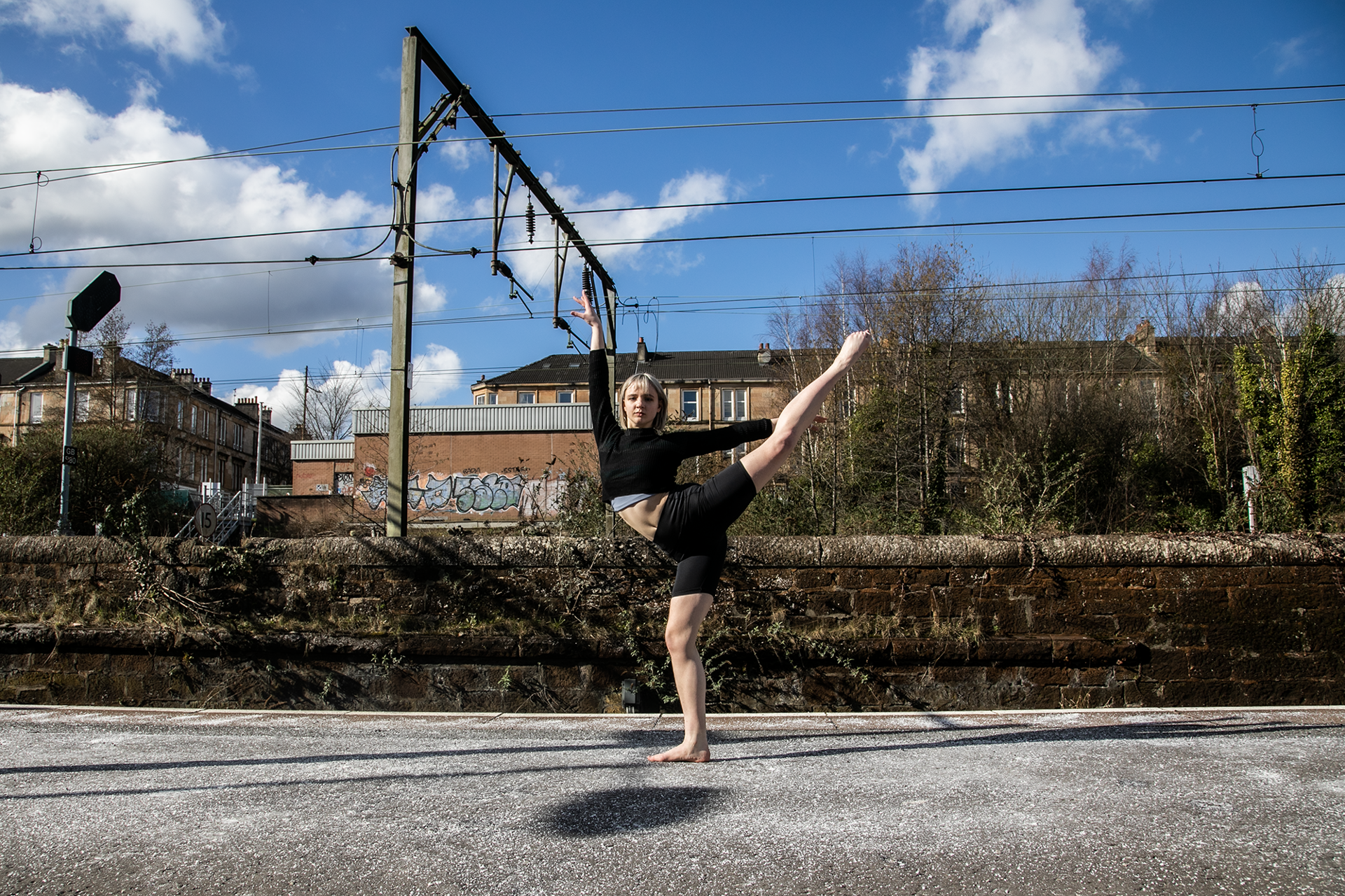 white female cropped blonde hair on a train platform with one leg kicked high to the side up, another on demi point and one arm above and another to the right. Wearing black shorts and black crop top with bare feet white female cropped blonde hair on a train platform with one leg kicked high to the side up, another on demi point and one arm above and another to the right. Wearing black shorts and black crop top with bare feet