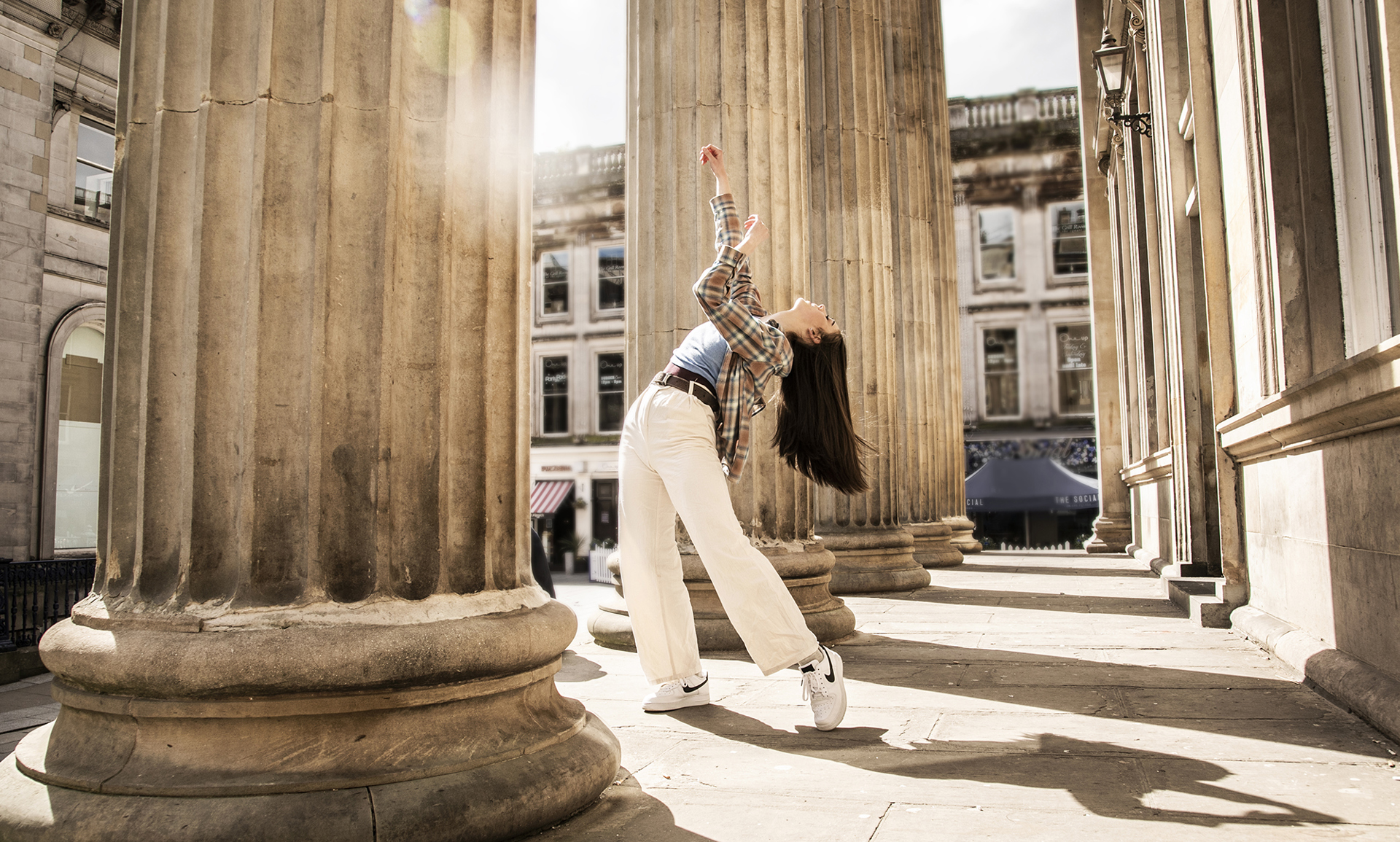 Young female dancer infront of Glasgow coucil building with large pillars. Legs lunched arms above the head. White female dance with long brown hair wearing checked shirt blue top and white trousers 