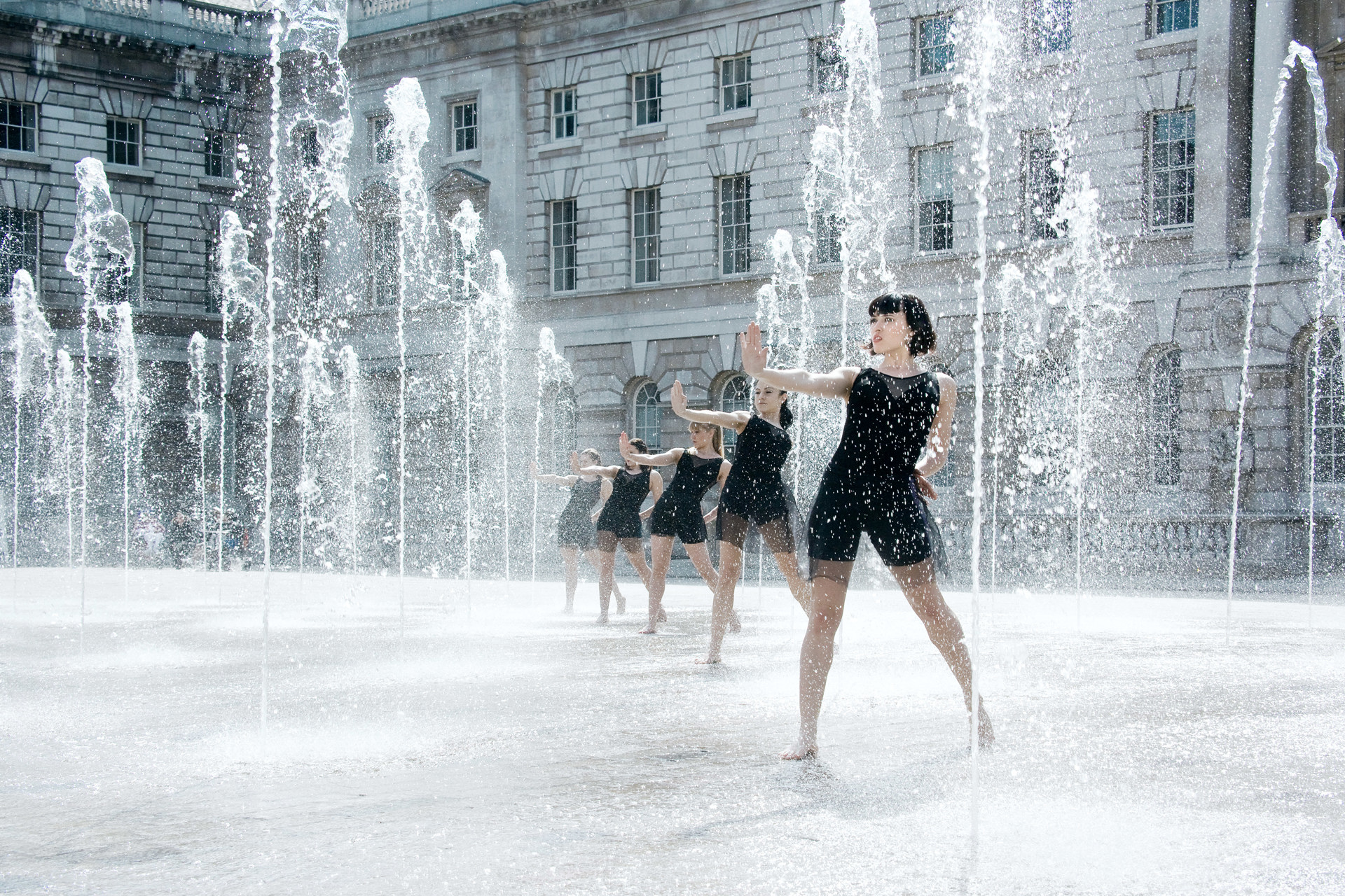 five female dancers with one arm reached out all standing in a water fountain. Wearing black outfits. five female dancers with one arm reached out all standing in a water fountain. Wearing black outfits.