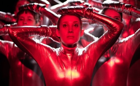 female with hands on head with other dancers in the background copying the movement. All wearing lycra bodysuit with red lighting. 